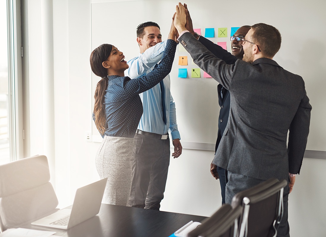 Employee Benefits - Four Cheerful Employees Standing in a Circle Putting Their Hands Together to Give Each Other High Fives While Standing in a Conference Room